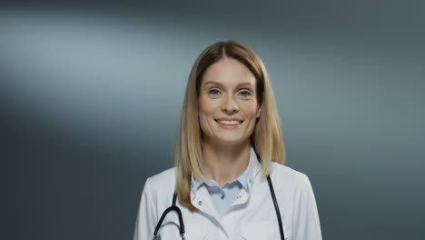 Portrait-shot-of-the-young-Caucasian-blonde-pretty-woman-physician-in-the-white-medical-gown-turning-to-the-camera-and-smiling-happily-on-the-gray-wall-background.-Close-up.